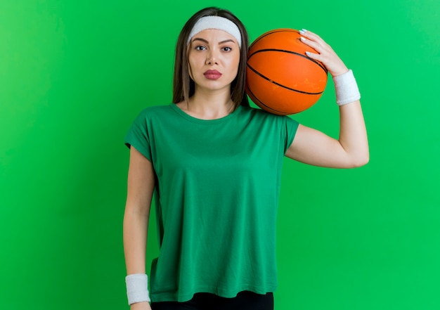 Mujer deportiva joven confiada con diadema y muñequeras sosteniendo una pelota de baloncesto en el hombro mirando