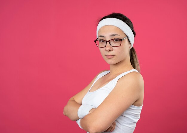 Mujer deportiva joven confiada en gafas ópticas con diadema y muñequeras se encuentra de lado con los brazos cruzados aislados en la pared rosa