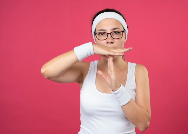 Mujer deportiva joven confiada en gafas ópticas con diadema y muñequeras gestos tiempo de espera signo aislado en la pared rosa