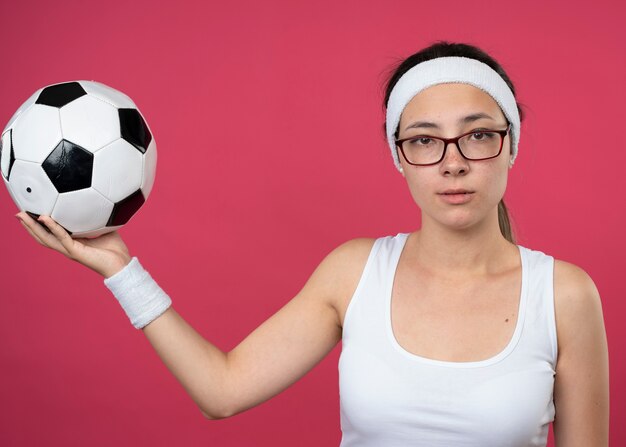 Mujer deportiva joven confiada en gafas ópticas con diadema y muñequeras sosteniendo la bola aislada en la pared rosa