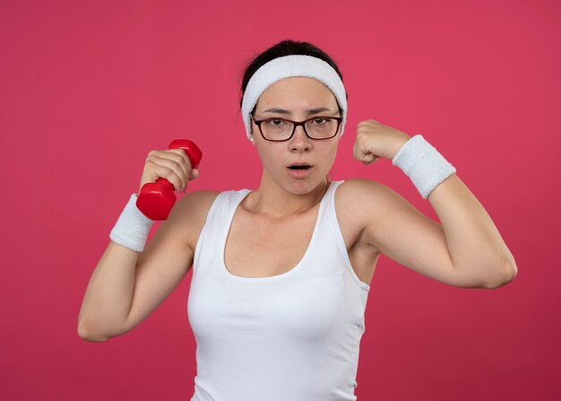 Mujer deportiva joven confiada en gafas ópticas con diadema y muñequeras sostiene mancuernas y tensa bíceps aislado en la pared rosa