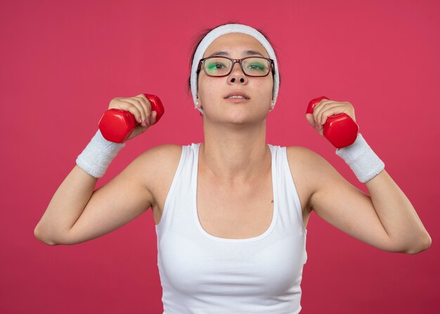 Mujer deportiva joven confiada en gafas ópticas con diadema y muñequeras sostiene pesas aisladas en la pared rosa