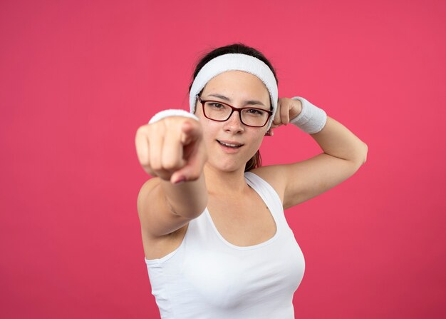 Mujer deportiva joven confiada en gafas ópticas con diadema y muñequeras tensa bíceps y puntos en el frente aislado en la pared rosa