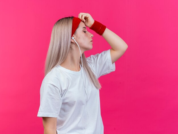 Mujer deportiva joven confiada con tirantes en los auriculares con diadema y muñequeras está de lado poniendo la mano en la frente aislada en la pared rosa