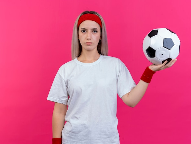 Mujer deportiva joven confiada con tirantes con diadema y muñequeras sostiene la bola aislada en la pared rosa