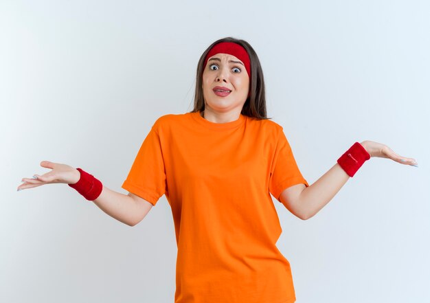 Foto gratuita mujer deportiva joven confundida con diadema y muñequeras mostrando las manos vacías mirando aislado