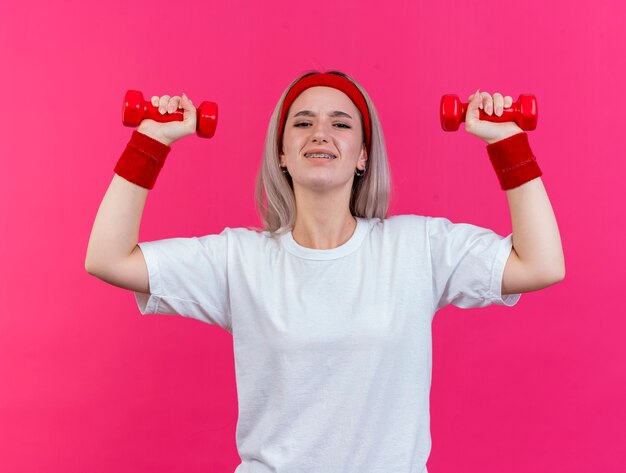 Mujer deportiva joven disgustada con tirantes con diadema y muñequeras se encuentra con las manos levantadas sosteniendo pesas aisladas en la pared rosa