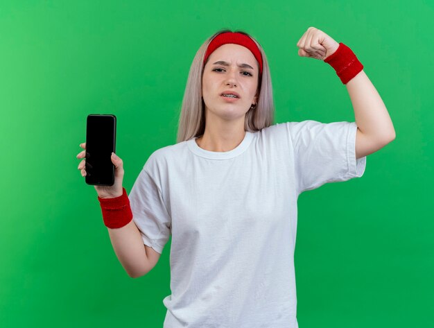 Mujer deportiva joven disgustada con tirantes con diadema y muñequeras se encuentra con el puño levantado y sostiene el teléfono aislado en la pared verde