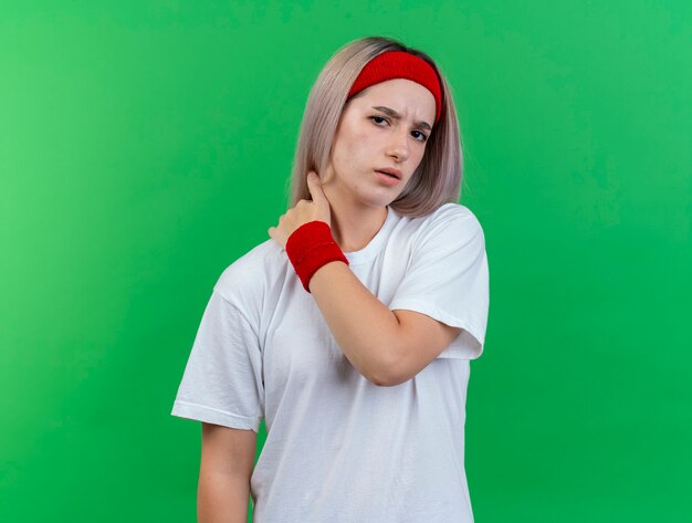 Mujer deportiva joven disgustada con tirantes con diadema y muñequeras pone la mano en el cuello aislado en la pared verde