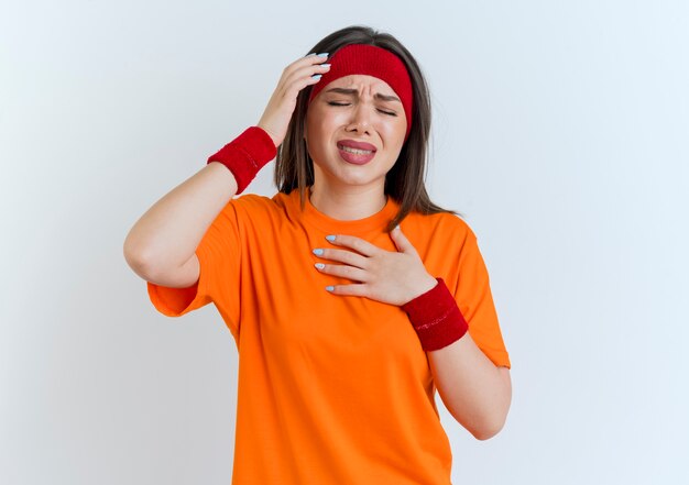 Mujer deportiva joven dolorida con diadema y muñequeras manteniendo las manos en la cabeza y en el pecho con los ojos cerrados