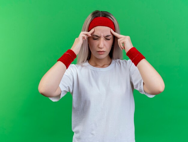 Mujer deportiva joven dolorida con tirantes con diadema y muñequeras pone los dedos en las sienes aisladas en la pared verde