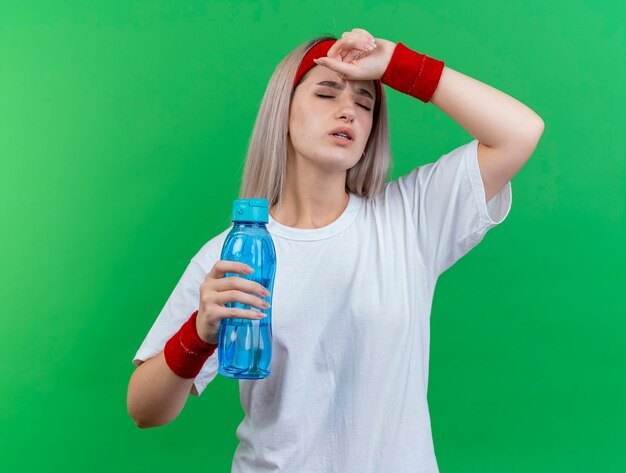 Mujer deportiva joven dolorida con tirantes con diadema y muñequeras pone la mano en la frente y sostiene la botella de agua aislada en la pared verde