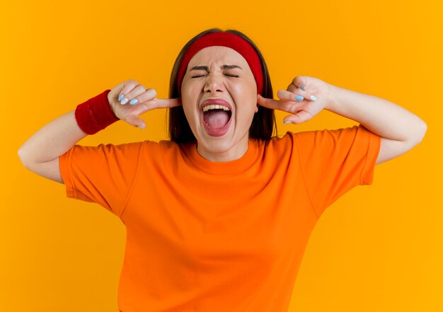 Mujer deportiva joven enojada con diadema y muñequeras manteniendo las manos en los oídos gritando con los ojos cerrados