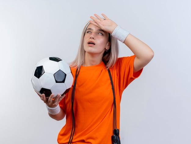 Mujer deportiva joven molesta con tirantes y con saltar la cuerda alrededor del cuello con diadema y muñequeras sostiene la pelota y pone la mano en la frente aislada en la pared blanca