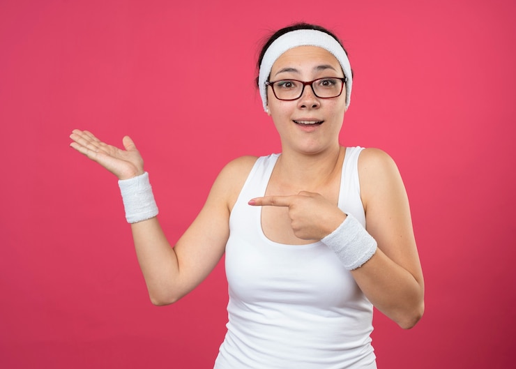 Foto gratuito mujer deportiva joven sorprendida en gafas ópticas con puntos de diadema y muñequeras en la mano vacía aislada en la pared rosa
