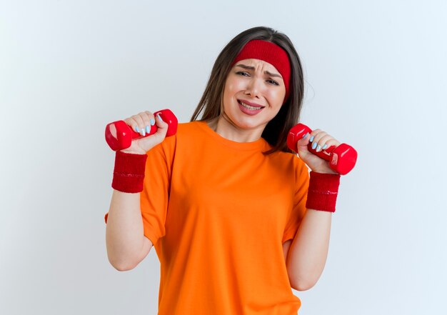 Mujer deportiva joven tensionada con diadema y muñequeras mirando sosteniendo pesas