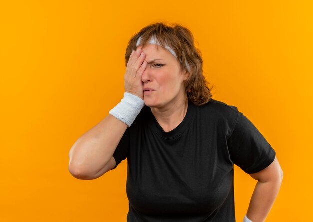 Mujer deportiva de mediana edad en camiseta negra con diadema con aspecto cansado cubriendo un ojo con el brazo parado sobre la pared naranja