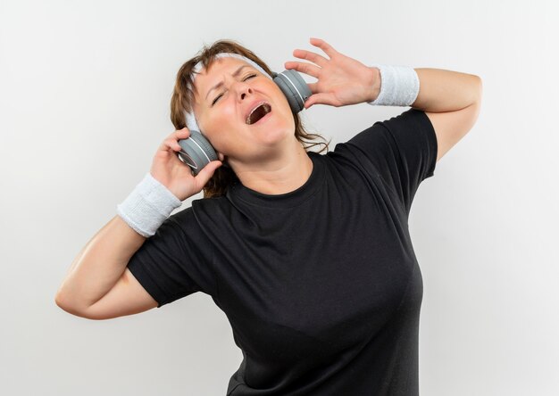 Mujer deportiva de mediana edad en camiseta negra con diadema y auriculares loco feliz disfrutando de la música de pie sobre la pared blanca