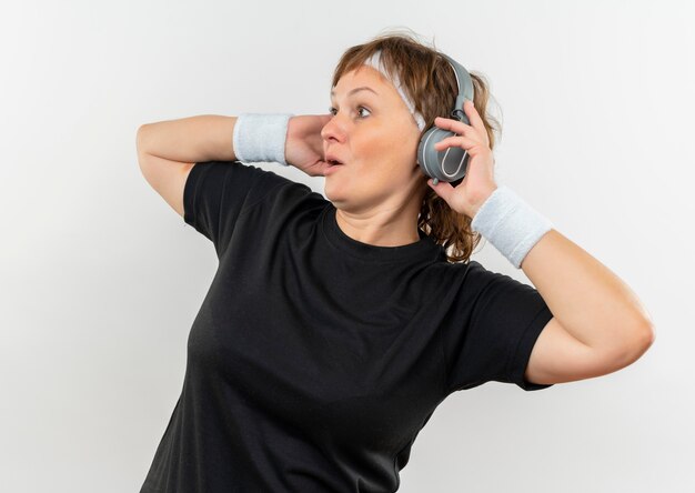 Mujer deportiva de mediana edad en camiseta negra con diadema y auriculares mirando sorprendido de pie sobre la pared blanca