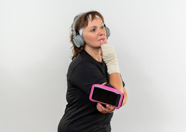 Mujer deportiva de mediana edad en camiseta negra con diadema y brazalete de teléfono inteligente mirando a un lado con expresión pensativa pensando de pie sobre la pared blanca