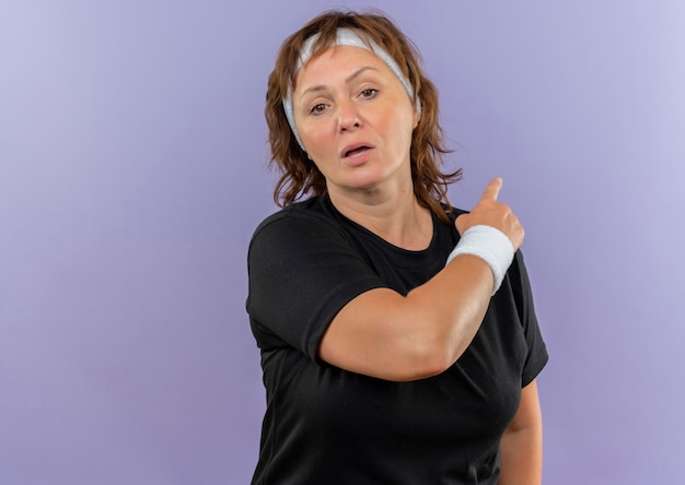 Mujer deportiva de mediana edad en camiseta negra con diadema cansado apuntando hacia atrás de pie sobre la pared azul