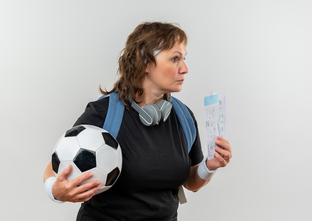 Mujer deportiva de mediana edad en camiseta negra con diadema y mochila sosteniendo boletos de avión y balón de fútbol mirando a un lado con expresión pensativa en la cara de pie sobre la pared blanca