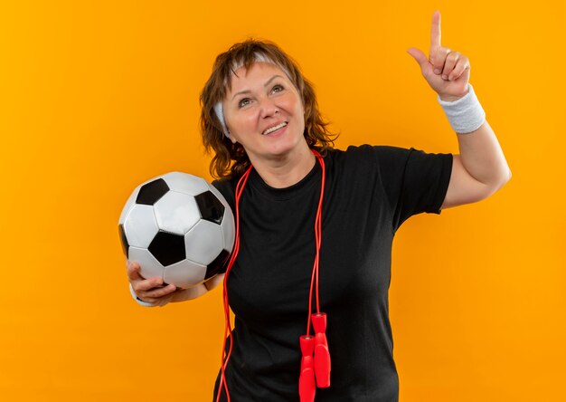 Mujer deportiva de mediana edad en camiseta negra con diadema sosteniendo un balón de fútbol apuntando con el dedo índice hacia arriba sonriendo de pie sobre la pared naranja