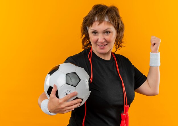 Mujer deportiva de mediana edad en camiseta negra con diadema sosteniendo un balón de fútbol levantando el puño feliz y positivo, concepto ganador de pie sobre la pared naranja