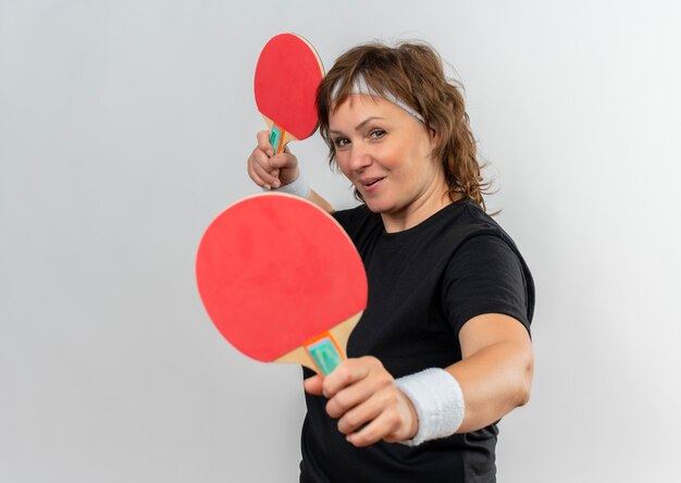 Mujer deportiva de mediana edad en camiseta negra con diadema sosteniendo dos raquetas de tenis de mesa con una sonrisa en la cara de pie sobre la pared blanca