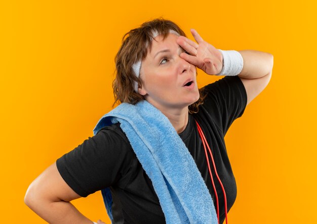 Mujer deportiva de mediana edad en camiseta negra con diadema y toalla en el hombro con aspecto cansado y agotado después del entrenamiento de pie sobre la pared naranja