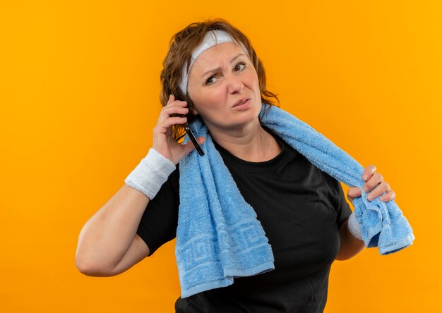 Mujer deportiva de mediana edad en camiseta negra con diadema y toalla en el hombro con aspecto cansado hablando por teléfono móvil de pie sobre la pared naranja