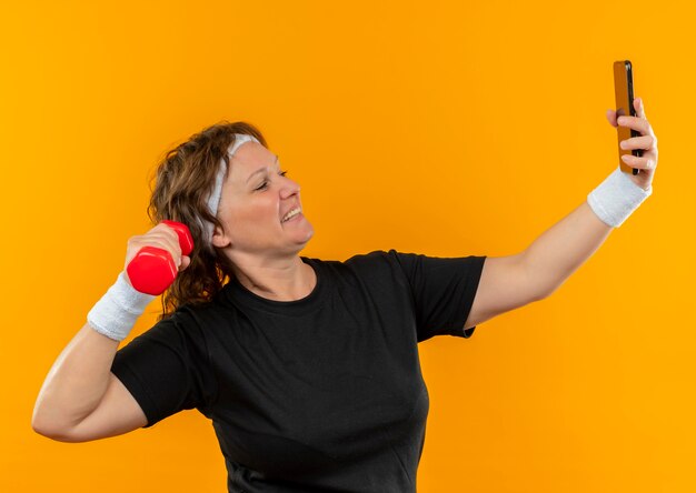 Mujer deportiva de mediana edad en camiseta negra con diadema trabajando con pesas de su teléfono inteligente tomando selfie de pie sobre la pared naranja