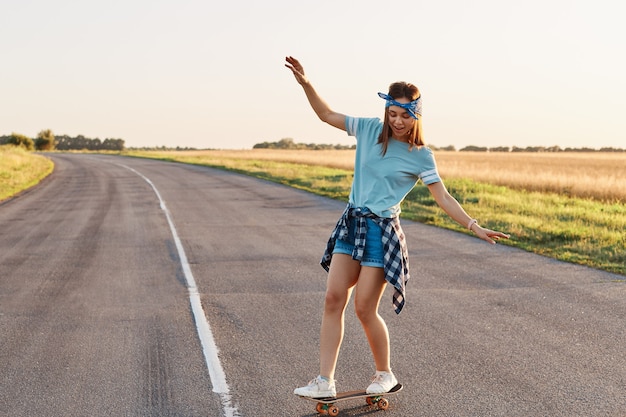 Mujer deportiva montando en la patineta en la carretera., Mujer deportiva delgada disfrutando del longboard, levantando las manos, con expresión concentrada feliz, estilo de vida saludable, espacio de copia.