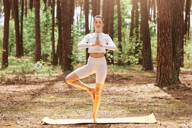 Mujer deportiva relajada de pie al aire libre con los ojos cerrados manteniendo las palmas juntas, de pie sobre una pierna, vistiendo ropa deportiva, disfrutando del entrenamiento en el hermoso bosque.