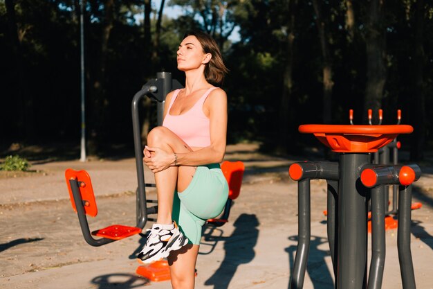 Mujer deportiva en ropa deportiva adecuada al atardecer en el campo de deportes estirando sus piernas antes después del entrenamiento