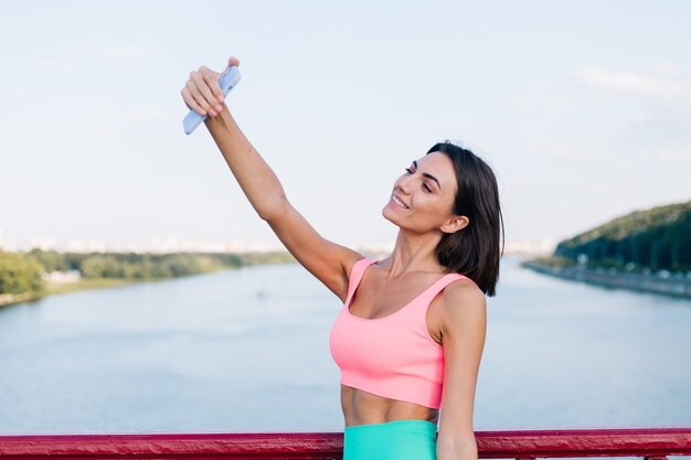 Mujer deportiva en ropa deportiva adecuada al atardecer en el puente moderno con vista al río feliz sonrisa positiva con teléfono móvil tomar foto video selfie para historias sociales