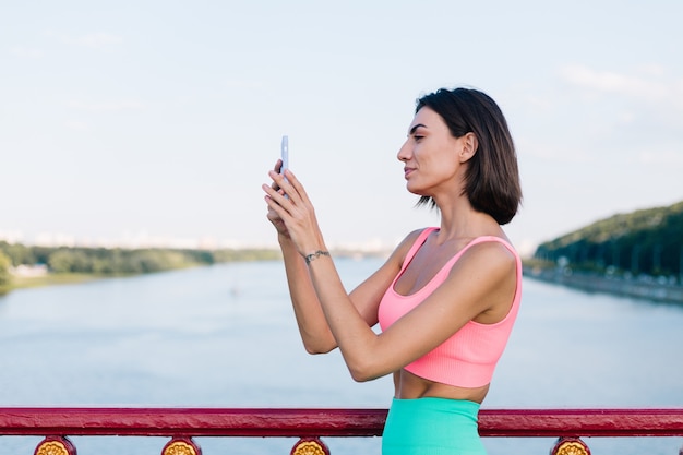 Mujer deportiva en ropa deportiva adecuada al atardecer en el puente moderno con vista al río feliz sonrisa positiva con teléfono móvil