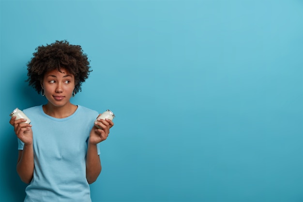 Mujer deportiva saludable con cabello afro sostiene frascos de vidrio de yogur fresco, va a preparar el desayuno, tiene una nutrición adecuada, se concentra a un lado, usa ropa informal, posa contra la pared azul