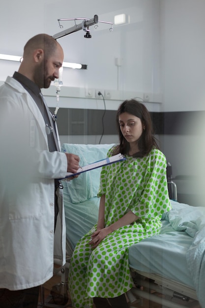 Foto gratuita una mujer deprimida en la cama del hospital descubre que tiene una enfermedad grave porque el médico sostiene un portapapeles con los resultados de las pruebas. paciente durante la consulta matutina sentado al lado de la cama sintiéndose triste después del tratamiento auditivo