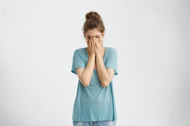 Mujer desanimada decepcionada con moño de pelo vistiendo una camiseta suelta azul que cubre su rostro con las manos cansadas y agotadas. Mujer desesperada que tiene depresión ocultando su rostro llorando con las manos