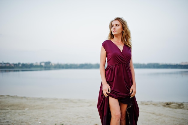 Mujer descalza sensual rubia en vestido rojo marsala posando contra el lago en la arena