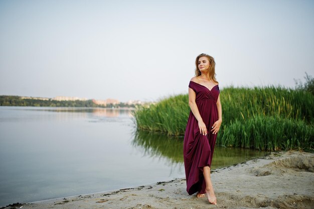 Mujer descalza sensual rubia en vestido rojo marsala posando contra el lago con juncos