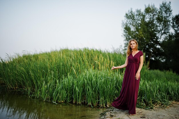 Mujer descalza sensual rubia en vestido rojo marsala posando contra el lago con juncos