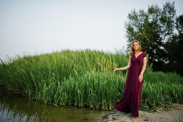 Mujer descalza sensual rubia en vestido rojo marsala posando contra el lago con juncos