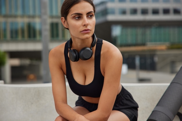 La mujer descansa después de la sesión de entrenamiento viste ropa deportiva tiene ejercicios regulares para trotar al aire libre contra la vista de la ciudad se toma un descanso