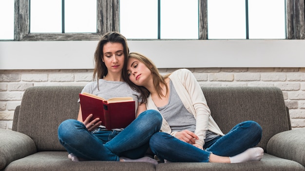 Mujer descansando en el hombro de su novia sentada en el sofá leyendo el libro