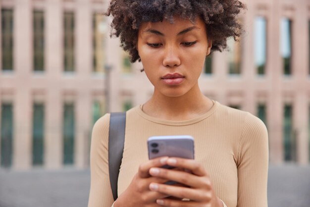 Mujer descarga la aplicación en el celular para controlar su actividad física vestida con ropa deportiva concentrada con expresión seria en la pantalla de poses de gadgets modernos al aire libre