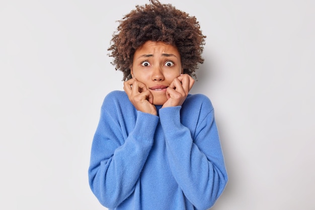 Mujer desconcertada ansiosa con el pelo rizado muerde las uñas de los dedos se encuentra insegura y nerviosa vestida con un jersey azul casual aislado sobre fondo blanco se siente incómoda. Concepto de reacciones humanas