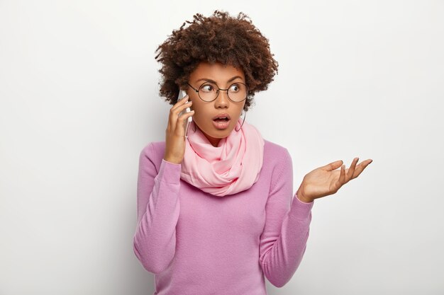 Mujer desconcertada con peinado afro, conversa con un colega o amigo, intenta resolver el problema a través de un teléfono inteligente, se concentra a un lado, usa lentes ópticos, suéter casual morado y bufanda alrededor del cuello