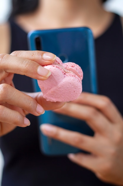 Mujer desenfocada tomando fotos de macaron en forma de corazón con teléfono inteligente